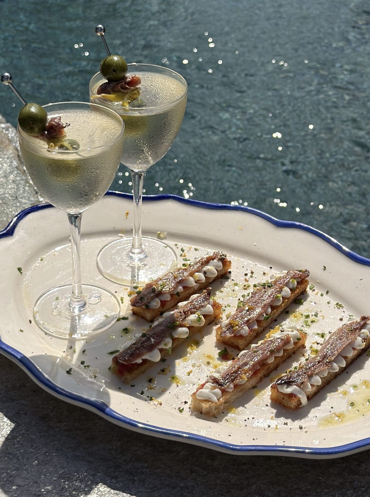 Two chilled Gilda martinis with olive, anchovy, and guindilla garnishes beside crisp anchovy toasties on a platter.