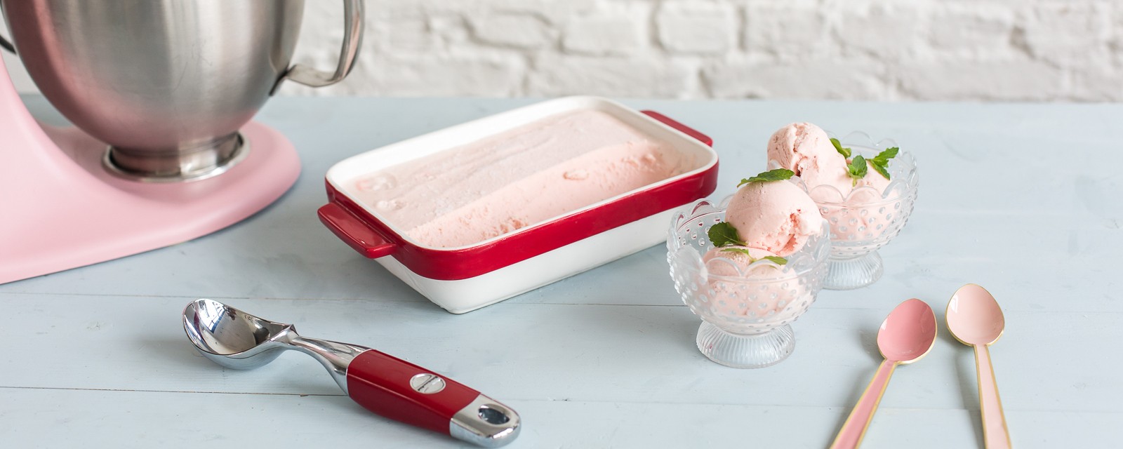 Pink strawberry ice cream in a red-rimmed dish and glass dessert bowls, with a stand mixer and scoops on a pale blue table.