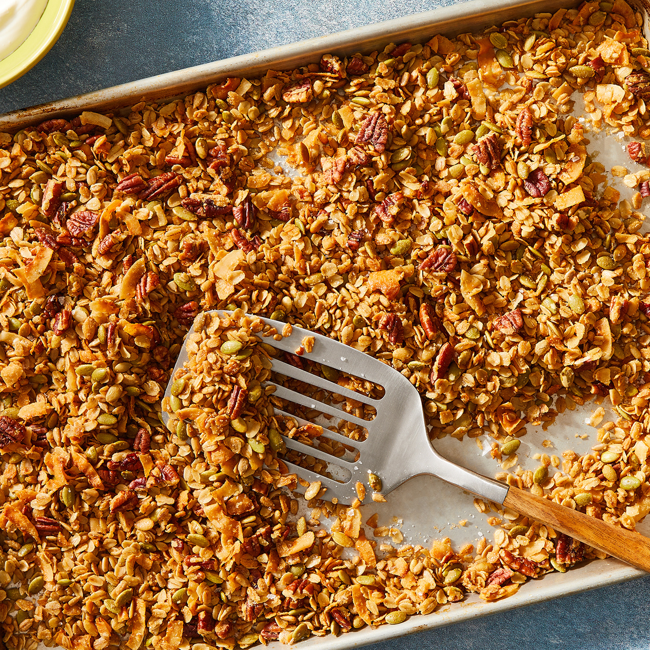 Golden granola with oats, nuts, seeds, and coconut spread on a baking sheet with a slotted spatula.