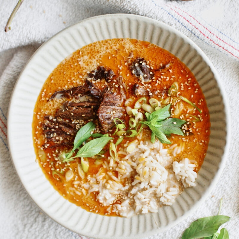A bowl of short rib coconut curry broth with rice, herbs, and sesame on a striped towel beside a spoon